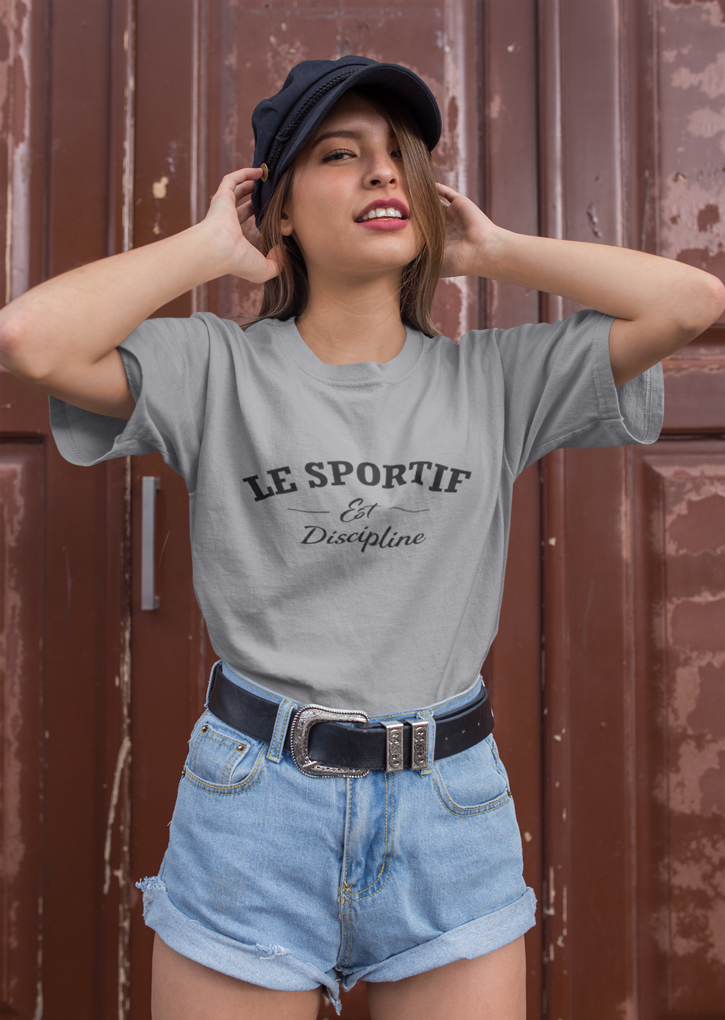 Person wearing a gray 'Le Sportif' t-shirt with denim shorts and a cap against a brown background